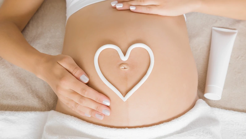 Young woman lying down on bed. Gentle hands touch. White heart shape created from cream on naked big belly. Care about perfect and smooth skin in pregnancy time. Close up.