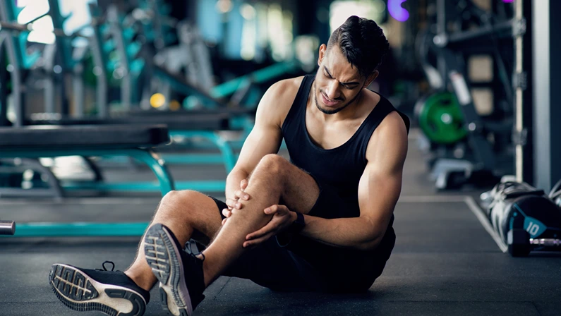 Sport Injury. Upset Arab Male Athlete Suffering From Knee Trauma While Training At Gym, Middle Eastern Man Sitting On Floor And Massaging Aching Area, Sporty Guy Frowning With Pain, Copy Space