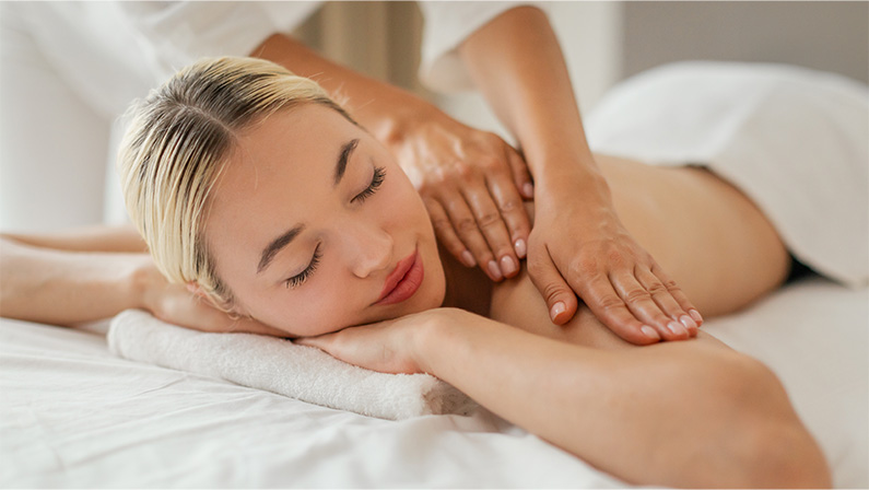 A woman is lying face down on a massage table while a massage therapist is applying pressure to her shoulders muscles. The woman appears relaxed and comfortable as she enjoys the soothing massage.