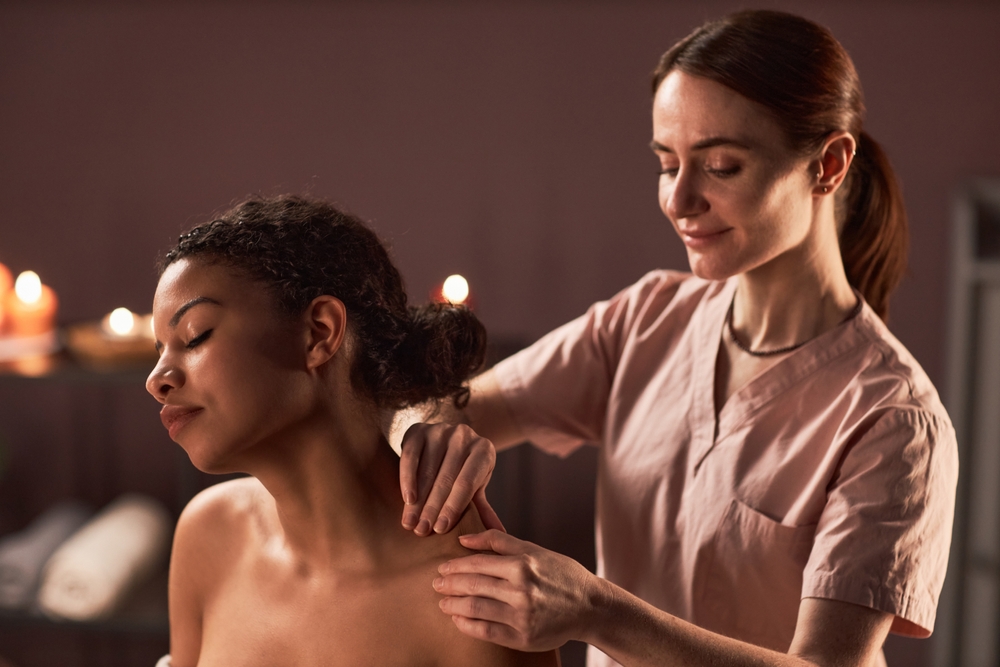 Smiling masseuse kneading shoulders of young woman